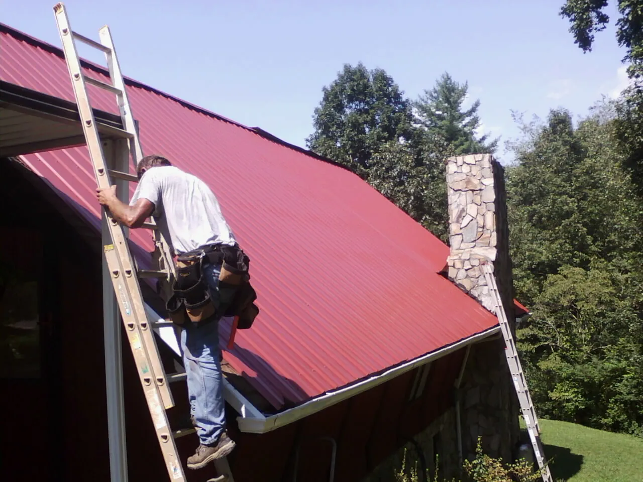 Roofer on a ladder beside a red metal roof and stone chimney — Soffit & Fascia Repair by Roofing Brothers in Middlefield